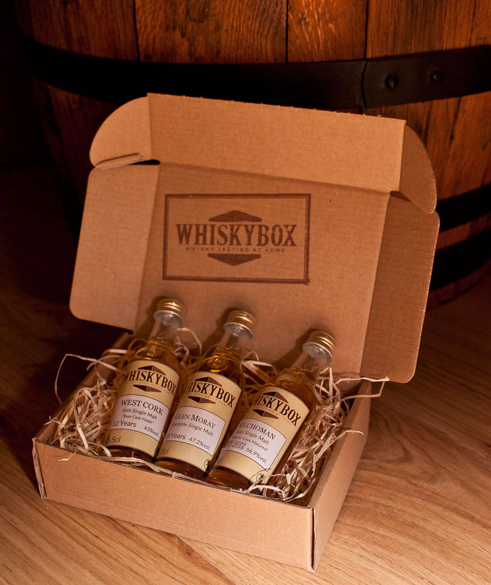 Flaschen der Whiskybox liegen im Karton vor einem Fass