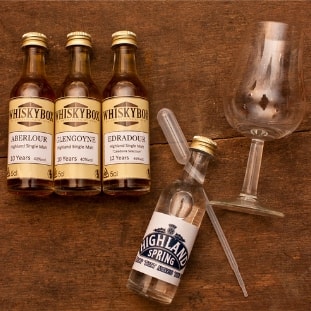 Flaschen, Pipette und Glas der Whiskybox - Whisky Tasting zu Hause - liegen auf einem Holzuntergrund.